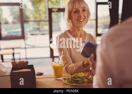 Il cliente soddisfatto. Affascinante Allegra donna senior dando una carta bancaria al barista e pagare per la sua insalata e succo in cafe mentre sorridente Foto Stock