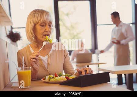 Cibo sano. Piacevole anziani donna seduta al cafe e insalata mangiare durante lo scorrimento verso il basso della timeline sul tablet e la verifica degli aggiornamenti dei suoi amici Foto Stock