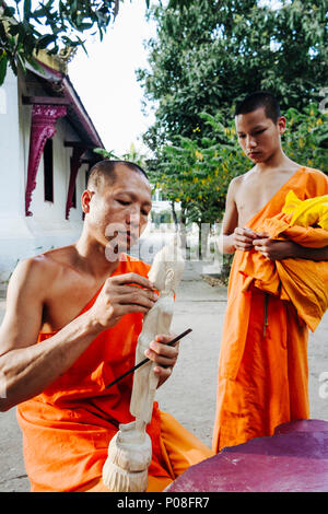 Luang Prabang, Laos, sud-est asiatico : un monaco buddista si ritaglia un di legno BUDDA STATUETTA come un giovane debuttante guarda, in un monastero a Luang Prabang. Foto Stock