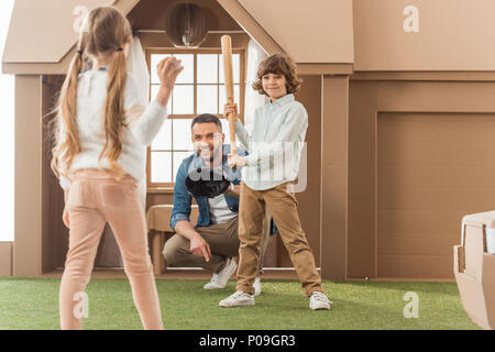 Padre insegnando ai bambini come a giocare a baseball nella parte anteriore della casa di cartone Foto Stock