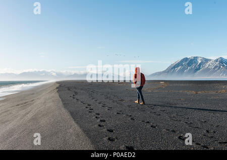 Uomo che cammina in riva al mare nella spiaggia di sabbia nera, lava spiaggia, montagne innevate, Hvalnes Riserva Naturale, a sud dell'Islanda Foto Stock