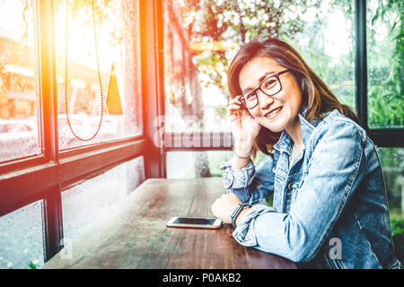 Occhiali nerd hipster donna asiatica seduta sorriso in finestre di vetro cafe con lo smartphone Foto Stock