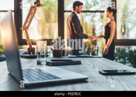 Scrivania in legno con laptop, smartphone e computer portatili collocati su di esso e giovani imprenditori si stringono la mano in background Foto Stock