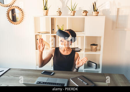 Giovane imprenditrice in abito nero seduto alla sua scrivania in ufficio, indossare occhiali per realtà virtuale Foto Stock