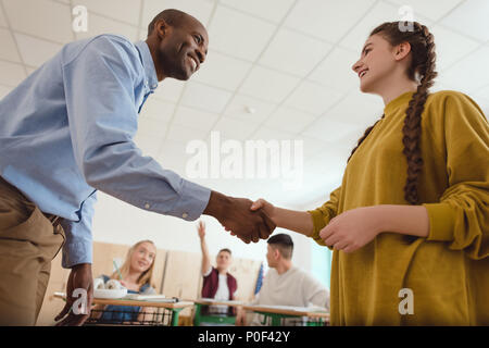 Basso angolo di vista sorridente americano africano insegnante agitando la mano di adolescenti schoolgirl con i compagni di classe seduta dietro in aula Foto Stock