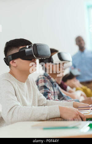 Vista laterale della società multietnica gli alunni delle scuola utilizzando la realtà virtuale degli auricolari e insegnante in piedi dietro Foto Stock