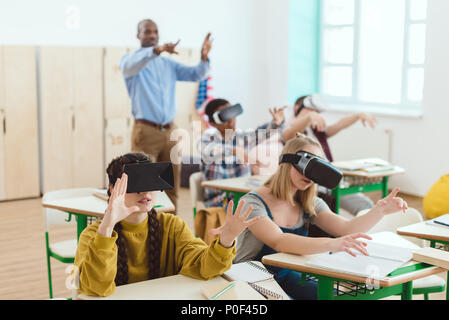 Alta scuola gli studenti adolescenti utilizzando la realtà virtuale degli auricolari e gesticolando insegnante in piedi dietro in aula Foto Stock