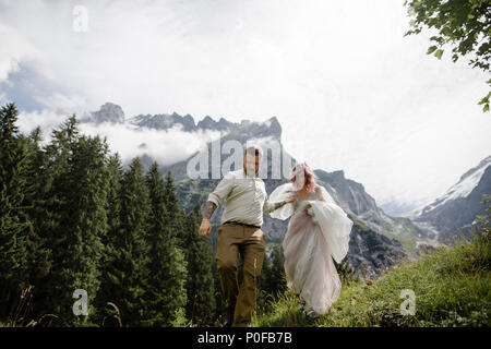 Giovane e bella sposa e lo sposo tenendo le mani e piedi sul verde prato di montagna nelle Alpi Foto Stock