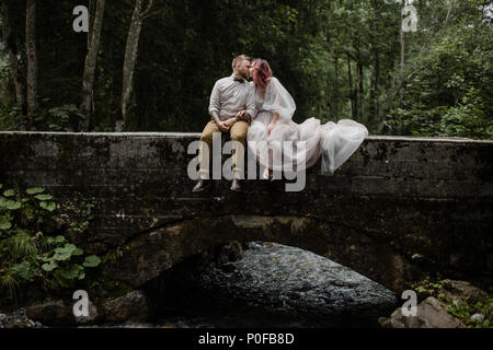 Bella giovani sposi kissing mentre è seduto sul ponte sopra il fiume di montagna nelle Alpi Foto Stock