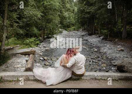 Vista posteriore del giovane e bella sposa e lo sposo seduto sul ponte sopra il fiume di montagna nelle Alpi Foto Stock