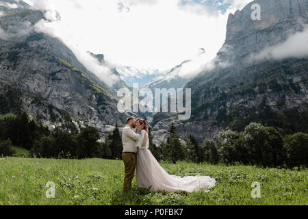 Felice sposa e lo sposo abbraccia e bacia sulla montagna verde prato con nuvole nelle Alpi Foto Stock