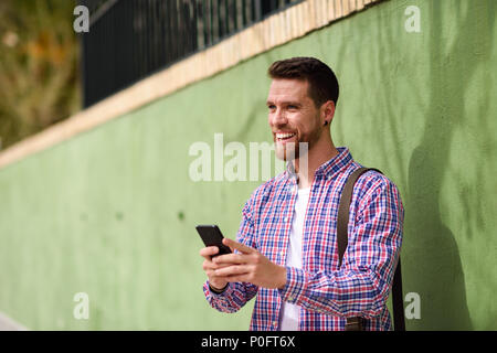 Giovane uomo a ridere con il suo smart phone in background urbano. Ragazzo indossa abiti casual. Il concetto di stile di vita. Foto Stock