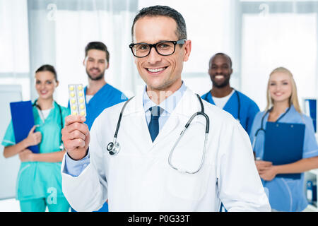 Bella coppia medico tenendo pillole con i colleghi in piedi insieme sullo sfondo Foto Stock
