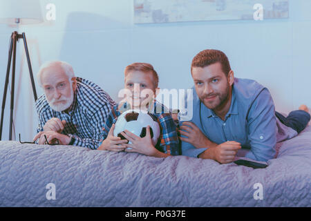 Little Boy tenendo palla calcio, suo padre e suo nonno giacente sul letto insieme e guardare la partita di calcio Foto Stock