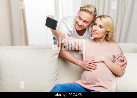 Felice la donna incinta con il marito tenendo selfie con lo smartphone a casa Foto Stock