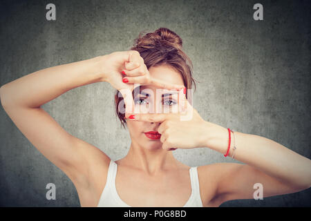 Ragazza giovane rendendo il telaio con le dita e guardando attraverso limiti a fotocamera su sfondo grigio Foto Stock