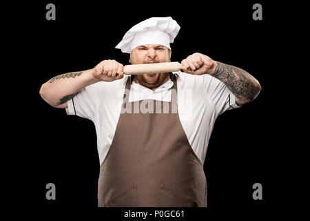 Arrabbiato giovane chef di hat e grembiule mordere mattarello isolato su nero Foto Stock