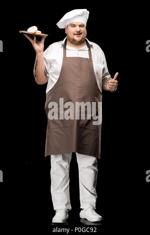 Allegro chef in grembiule e hat holding ciambelle sulla piastra e mostrando il pollice in alto Foto Stock