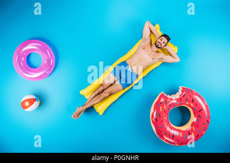 Vista superiore del bel giovane uomo in pantaloncini corti in appoggio sul materasso gonfiabile e sorridente in telecamera su blu Foto Stock