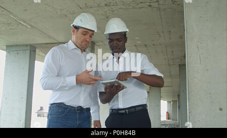 African American ingegnere e architetto caucasine utilizzando tavoletta digitale e indossare i caschi di sicurezza al sito in costruzione Foto Stock