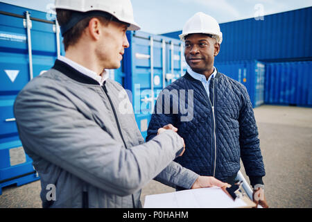 Due ingegneri sorridente indossando hardhats agitando mani insieme mentre in piedi da contenitori di spedizione in uno scalo merci Foto Stock