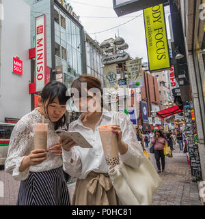 Due giovani donne, tenendo bicchieri per bevande, godetevi la lettura di un messaggio di testo sul telefono cellulare, Shibuya street, Tokyo, Giappone Foto Stock