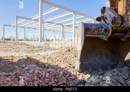 Escavatore attrezzo, benna, lama, alto edificio di cemento è dietro al sito di costruzione. Foto Stock