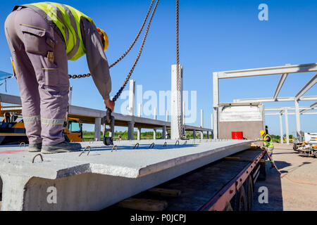 Lavoratore sta preparando il gancio della gru per lo scarico di un travetto in cemento da camion rimorchio. Foto Stock