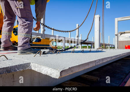 Lavoratore sta preparando il gancio della gru per lo scarico di un travetto in cemento da camion rimorchio. Foto Stock