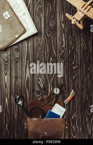 Vista superiore della disposizione del giocattolo di legno piano, borsa con lenti di ingrandimento, coltello da tasca, passaporto, biglietto, occhiali e bussola sul legno scuro tablet Foto Stock