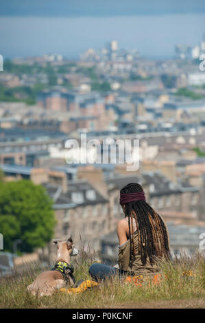 Molto cool, bohemian girl siede nell'erba lunga sulla Carlton Hill in una giornata di sole con Edimburgo al di sotto di lei e il suo cane Foto Stock