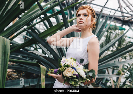 Bella giovane i capelli rossi sposa azienda bouquet nuziali e guardando lontano nel giardino botanico Foto Stock