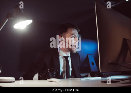 Agente emotivo con cuffia urlando mentre guardando lo schermo del computer nel buio Foto Stock