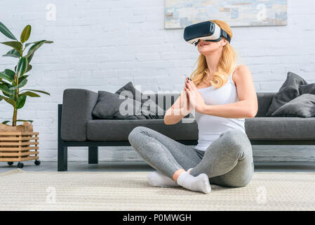 Giovane donna a praticare yoga nella posizione del loto e utilizzando la realtà virtuale auricolare Foto Stock