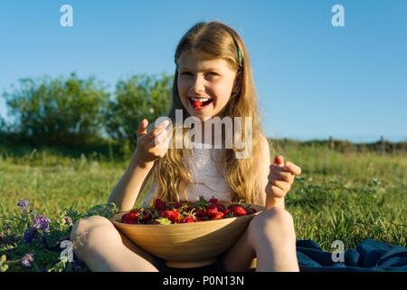 Attraente ragazza bambino mangiare fragole. Natura, sfondo verde prato, in stile country. Foto Stock