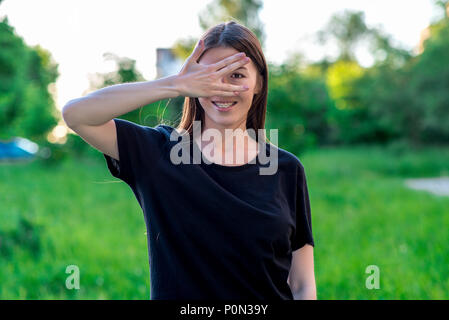 La bruna ragazza sorride felicemente. Dehors estivo in aria fresca. Gesto della mano e mostra che essi sono sbirciando attraverso le dita. Le bretelle sono sui denti. Foto Stock