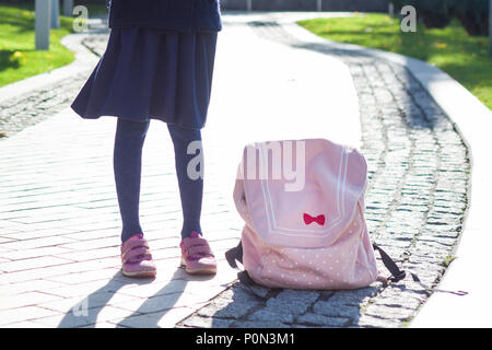 Scuola, scolari. Foto di una ragazza per le gambe in un mantello scuro con un zaino rosa sul marciapiede. Foto Stock