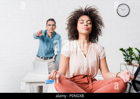 Giovane imprenditrice rilassante e meditando in ufficio con un collega di sesso maschile dietro Foto Stock