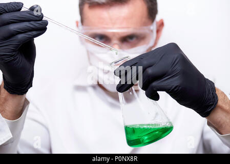 Un tubo di prova con una goccia e un pallone con un liquido verde nelle mani di uno scienziato in occhiali, maschera e guanti neri. Un chimico sta conducendo un ex Foto Stock