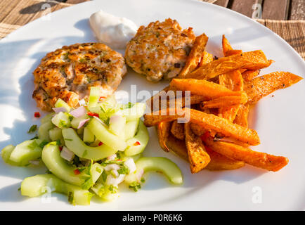 Close up White dinner piastra con il sud-est asiatico il cibo servito nel sole. Salmone fishcakes, insalata di cetrioli e patate dolci chips Foto Stock