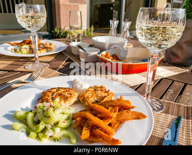 Ristoranti sudest asiatici servita sul patio esterno tabella nella luce del sole con cristallo bicchieri da vino riempito di vino bianco. Salmone fishcakes, insalata di cetrioli e patate dolci chips Foto Stock