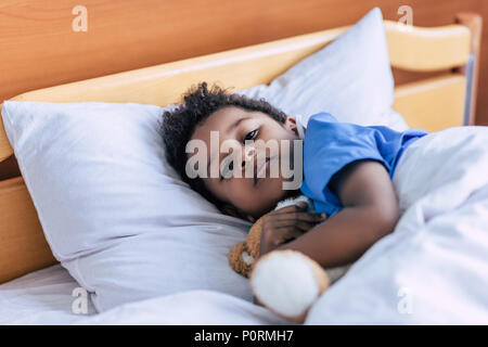 Ritratto di African American boy abbracciando Teddy bear mentre giaceva a letto in clinica Foto Stock