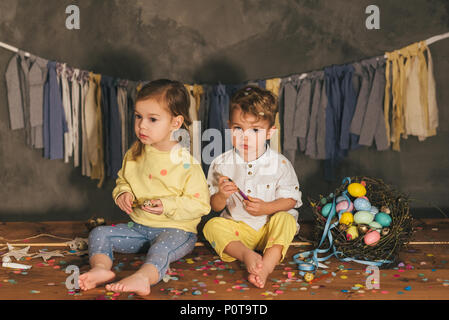 Adorabili poco i bambini seduti su rusitc pavimento in legno con cesto di uova di pasqua e coriandoli Foto Stock