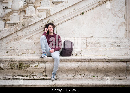 Bel giovane uomo ascoltare musica con le cuffie sulla sua testa. Giovane uomo rilassante nella parte anteriore del muro di pietra Foto Stock