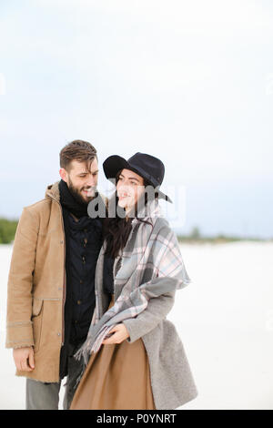 Felice l'uomo e la donna che indossa cappotti e sciarpe in piedi nella neve invernale dello sfondo. Foto Stock