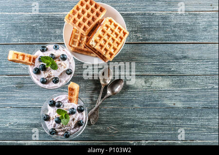 Ricotta dolce con i mirtilli in un bicchiere di vetro e in Casa Viennese di cialde su uno sfondo di legno. Vista dall'alto. Spazio di copia Foto Stock