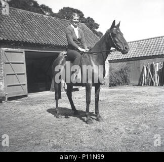 1951, storico, giovane donna seduta su un cavallo al di fuori di una stabile in un cortile, Inghilterra, Regno Unito. Foto Stock