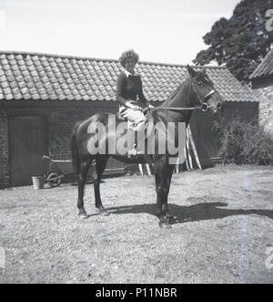 1951, storico, giovane donna seduta su un cavallo al di fuori di una stabile in un cortile, Inghilterra, Regno Unito. Foto Stock