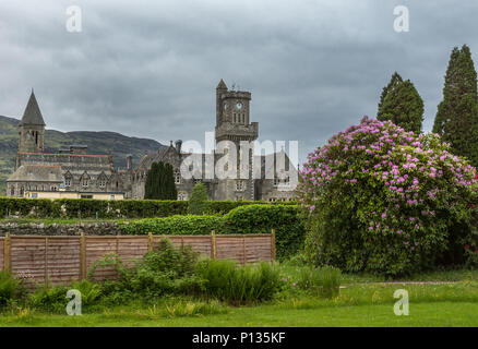 Fort Augustus, Scozia - Giugno 11, 2012: Abbey Highland Club con la torre dell orologio fronteggiata da giardino con fiori di colore rosa bush sotto pesante cloudscape. Le colline sulla Foto Stock