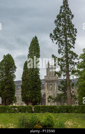 Fort Augustus, Scozia - Giugno 11, 2012: la torre dell orologio di Abbey Highland Club, visto tra gli alberi, fronteggiata da giardino sotto pesante cloudscape. Colline o Foto Stock